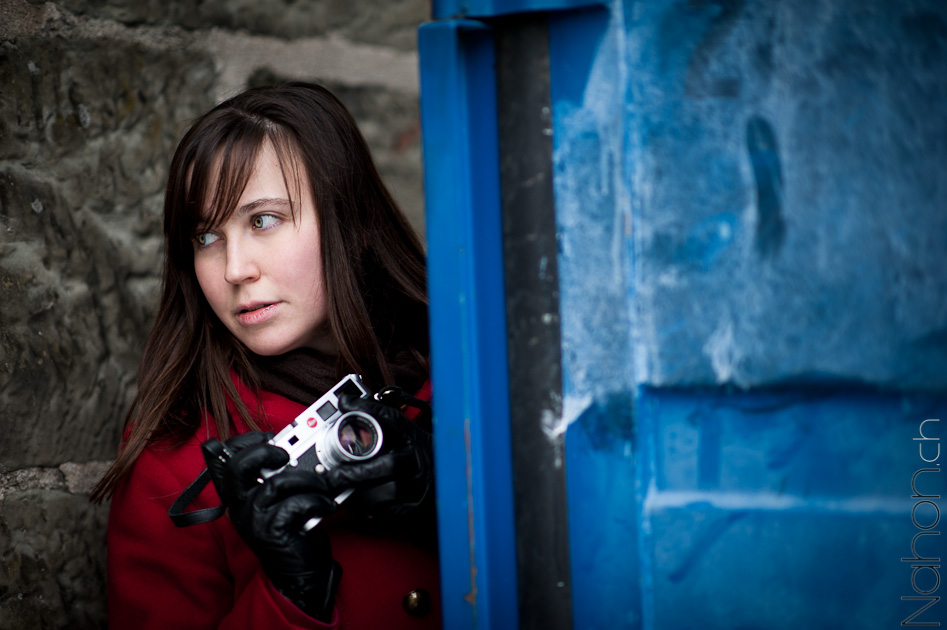 Photo d'un portrait d'une jeune femme en manteau rouge derrière un container bleu tenant un Leica M7