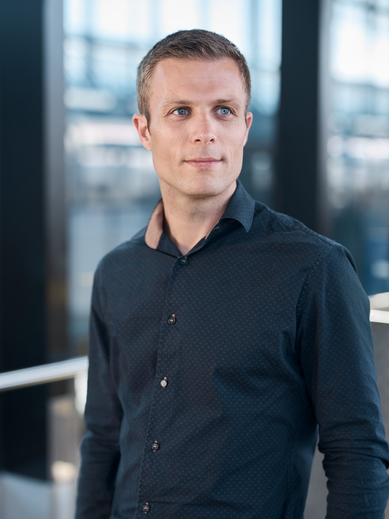 Portrait d'un homme professionnel au regard concentré, portant une chemise sombre à motifs, debout dans un environnement de bureau moderne éclairé par la lumière naturelle